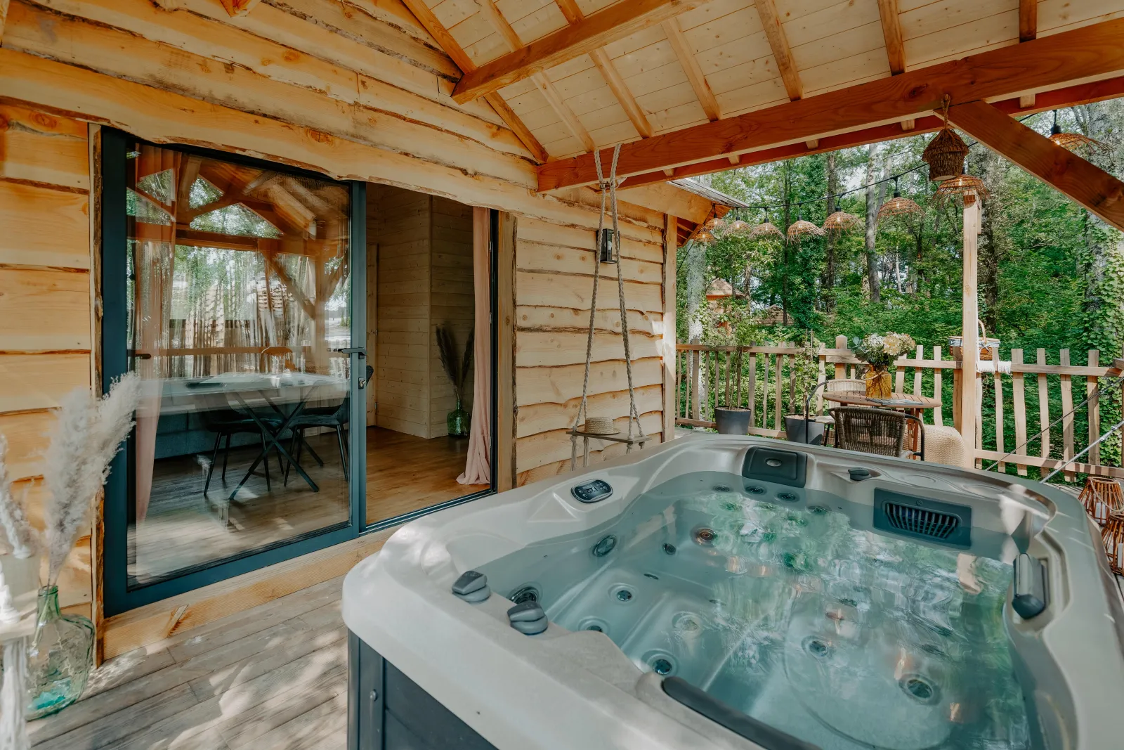 Covered terrace of a Girondine cabin featuring a hot tub, open glass doors leading to wooden interior, surrounded by lush greenery and forest