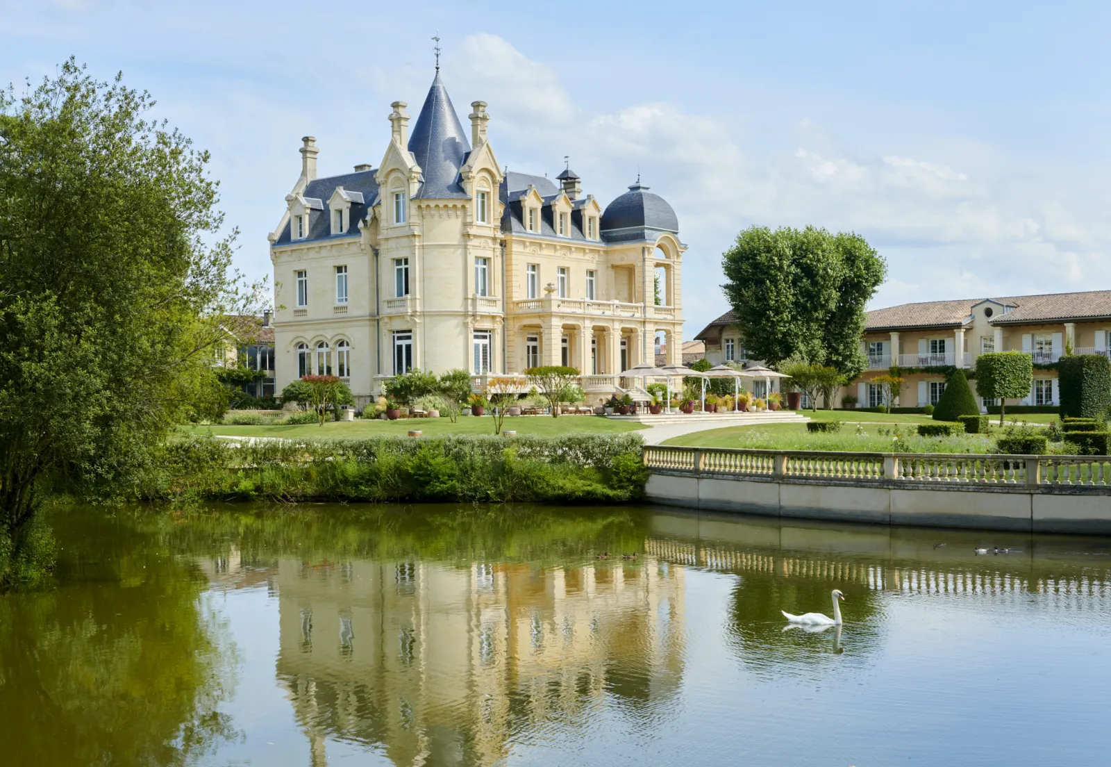 Grand Barrail Château Hotel in Bordeaux, a beige Second Empire castle with turrets reflected in a pond, surrounded by manicured gardens and outbuildings.