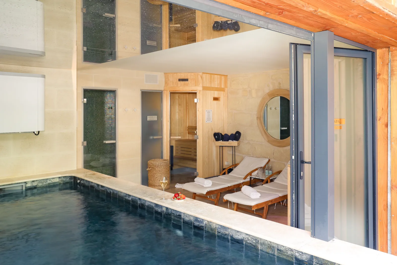 Indoor spa facility featuring a turquoise swimming pool, wooden lounge chairs with white cushions, wooden sauna, round mirror, and sliding glass doors, with warm beige and wood tones throughout.