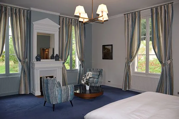 Elegant bedroom at Château Bellefont-Belcier in Bordeaux featuring a white bed, classical fireplace, blue armchairs, gray and gold curtains, bronze chandelier, and large windows overlooking the grounds.