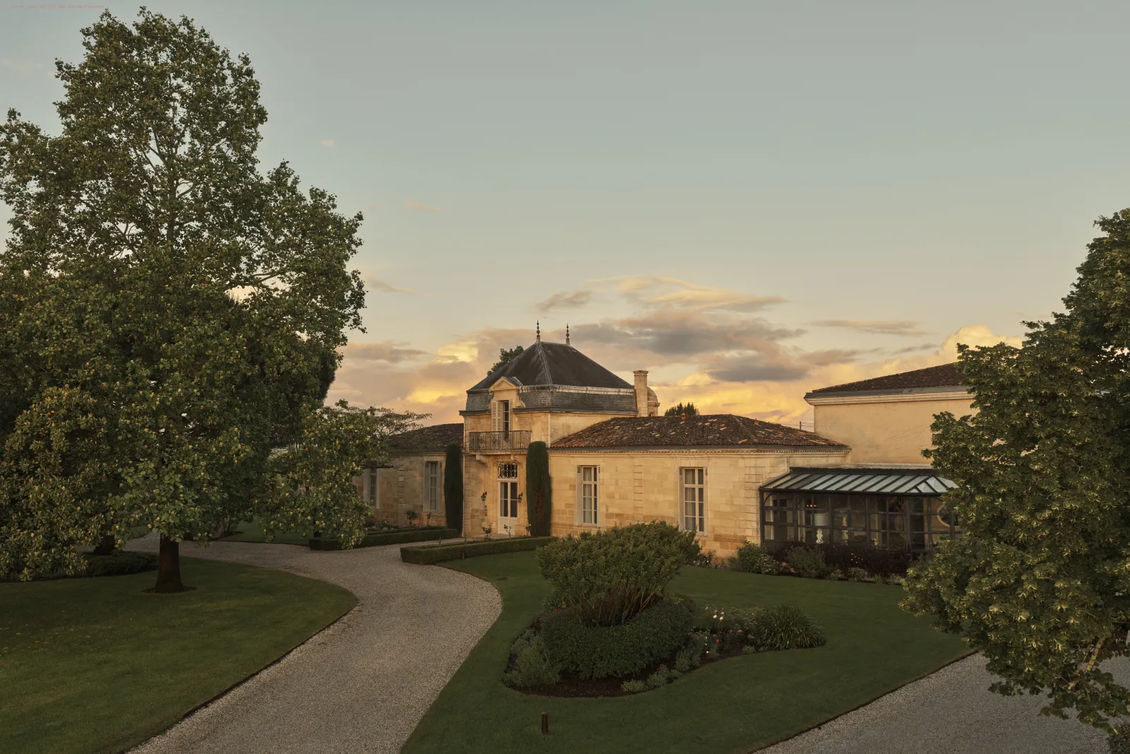 Sunset view of Château Cordeillan-Bages in Bordeaux, featuring its distinctive pale stone façade, mansard roof, surrounded by manicured gardens and mature trees.