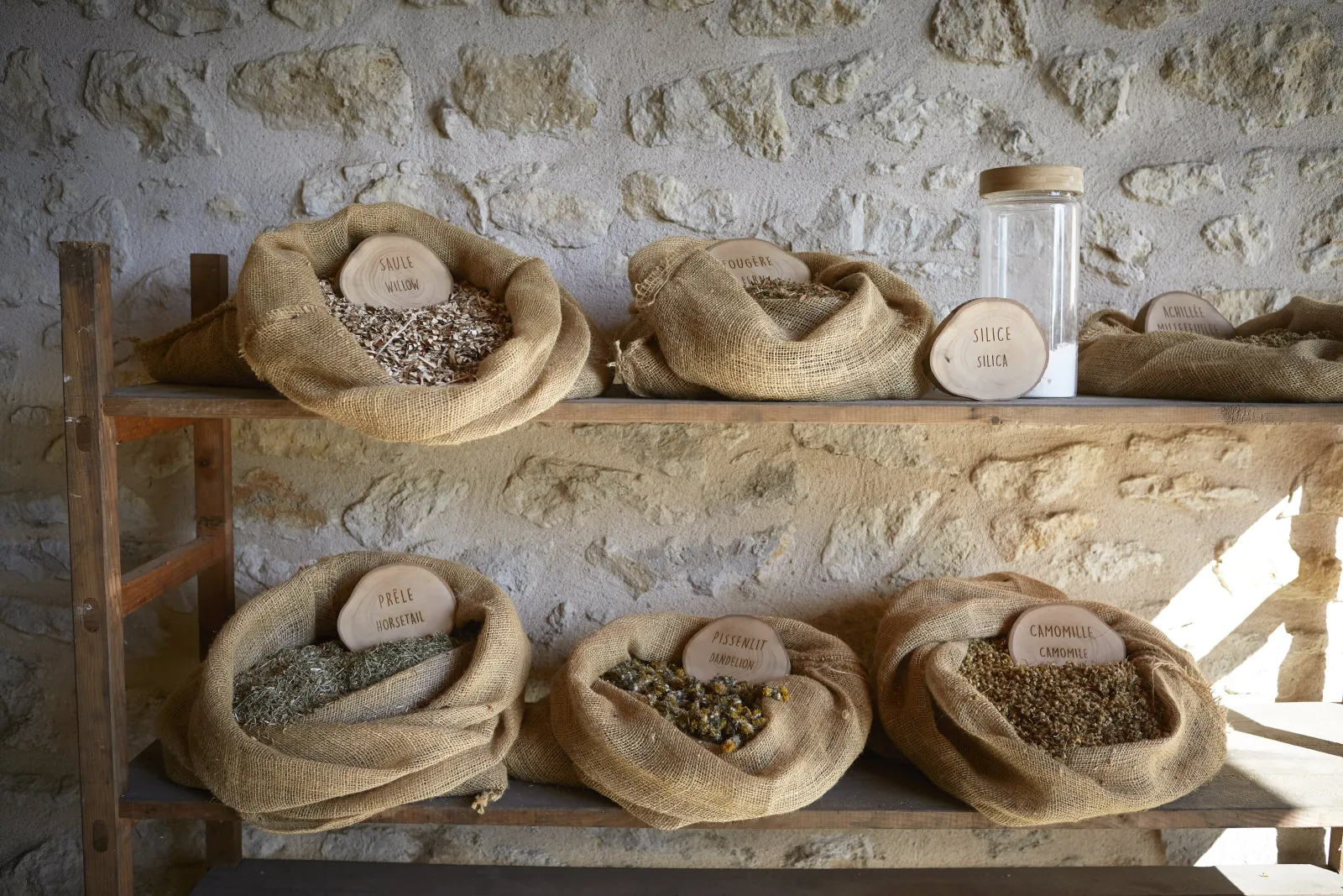 Stone shelves displaying burlap sacks filled with dried herbs and spices with handwritten labels, set against a whitewashed stone wall at Château de La Dauphine in Bordeaux.