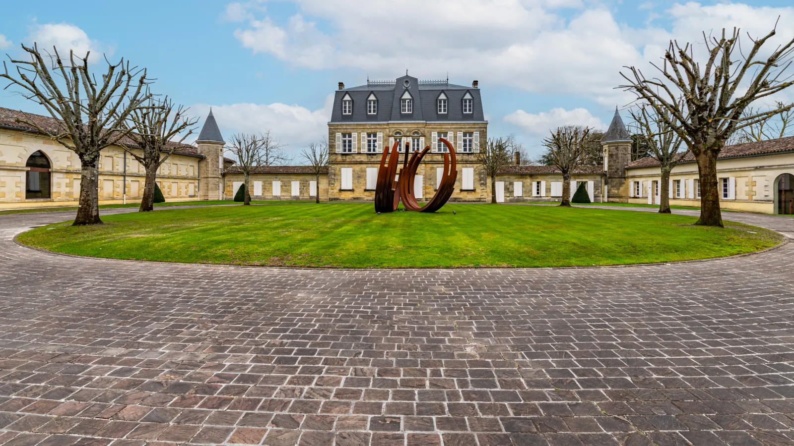 Château Malescasse in Bordeaux featuring a stone façade with mansard roof, surrounded by outbuildings, a circular paved courtyard, manicured lawn, and a modern red metal sculpture.