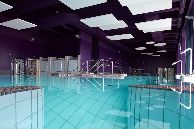 Modern indoor swimming pool featuring a turquoise basin, purple ceiling with LED lighting, metal access stairs, and white water slide in the center of La Calinésie aquatic center in Libourne.