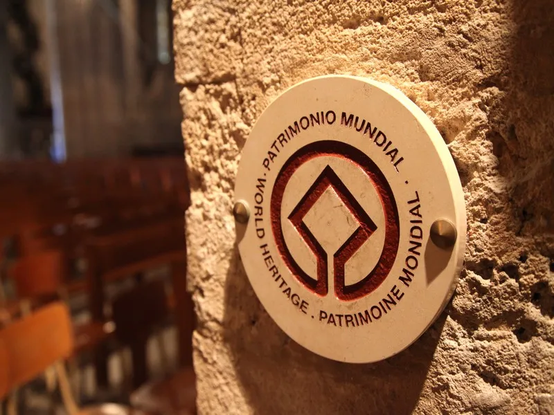 Circular UNESCO World Heritage plaque mounted on a stone wall, featuring a stylized diamond symbol and multilingual text indicating world heritage status.