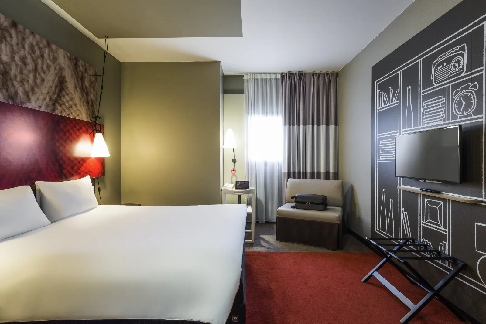 Modern hotel room featuring a white bed, yellow-green and burgundy walls, flat-screen TV, beige armchair, gray curtains and red carpet at Ibis Bordeaux Centre Gare Saint-Jean