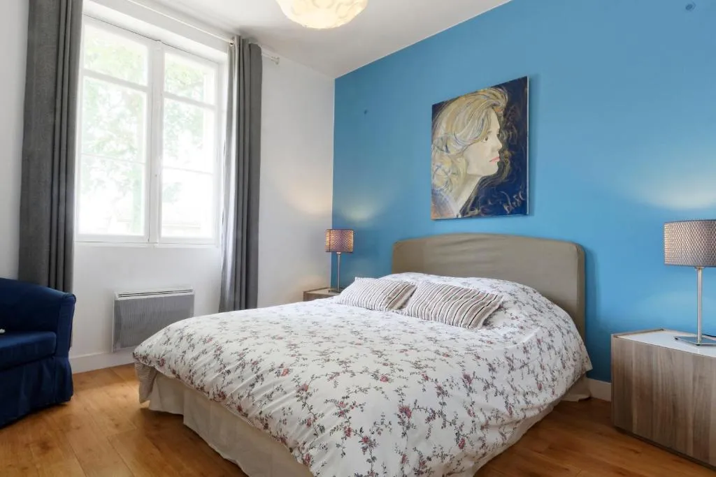 Modern bedroom at La Bastide de Siran, Bordeaux, featuring blue walls, queen-size bed with floral bedding, large windows overlooking greenery, wooden flooring and contemporary furniture