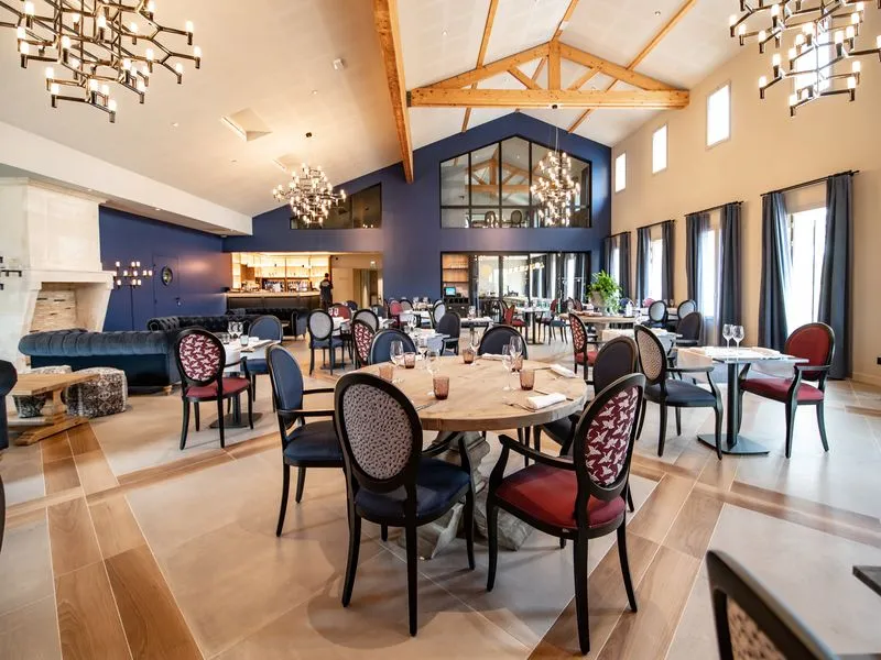 Elegant dining room at Château Fage La Maison des Vignes in Bordeaux, featuring round wooden tables, black and red patterned chairs, cathedral ceiling with exposed beams, modern chandelier, and large windows.
