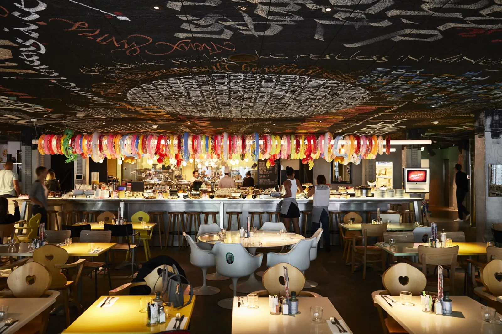 Interior of Mama Shelter restaurant in Bordeaux featuring colorful hanging lanterns from the ceiling, low tables, white chairs, and a bar counter in the background with warm modern lighting.