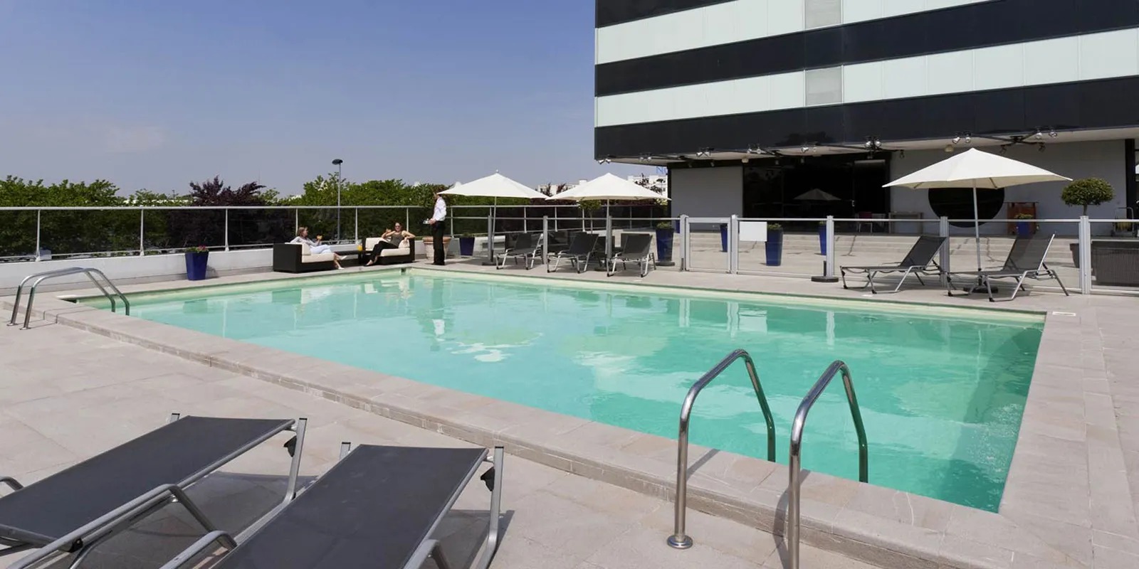 Outdoor swimming pool at Pullman Bordeaux Lac featuring turquoise water, sun loungers and umbrellas, modern terrace with black and white striped building in the background.