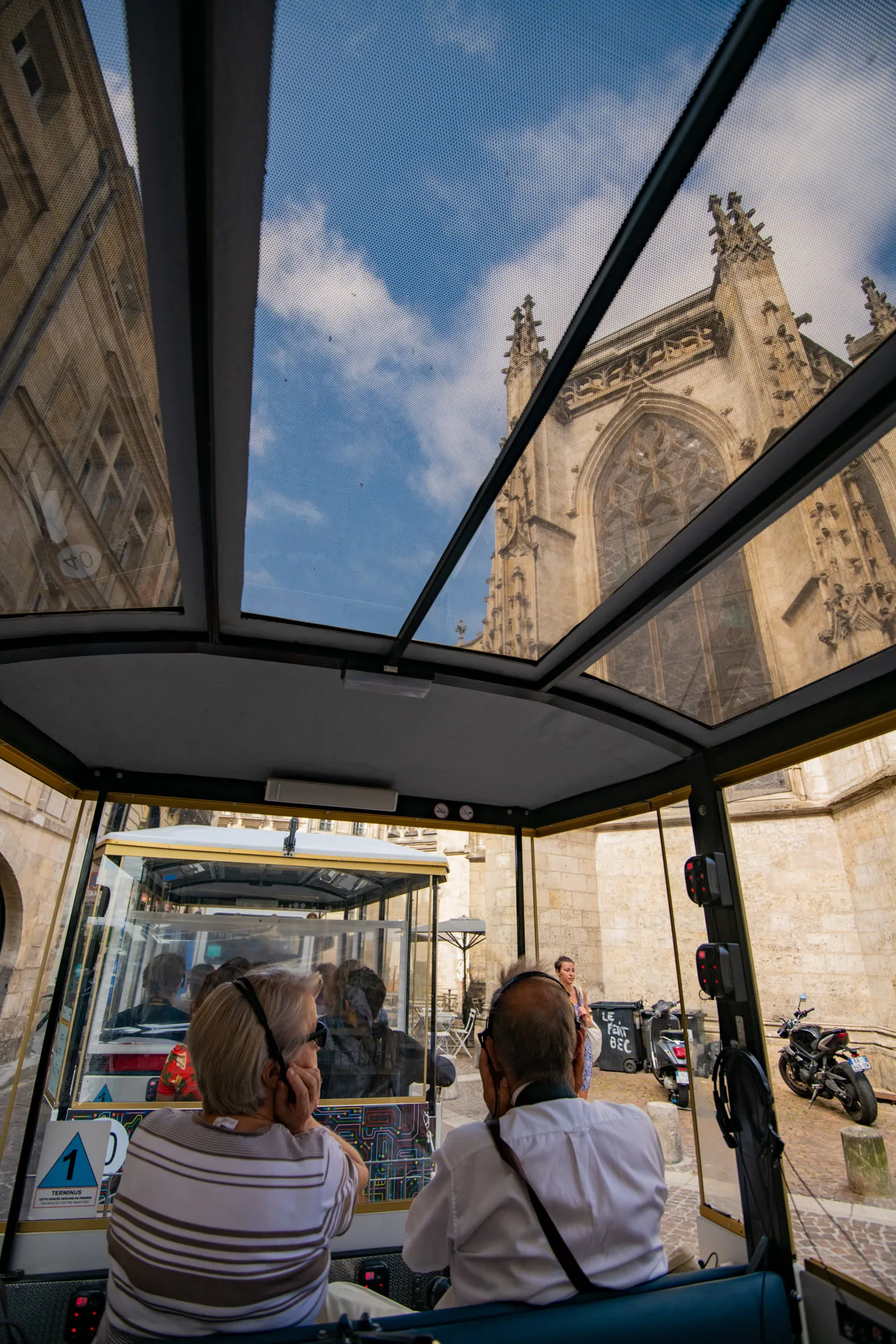 Interior view of an electric tourist train in Bordeaux with passengers seated, showing through panoramic windows the Gothic facade of a cathedral and urban architecture.