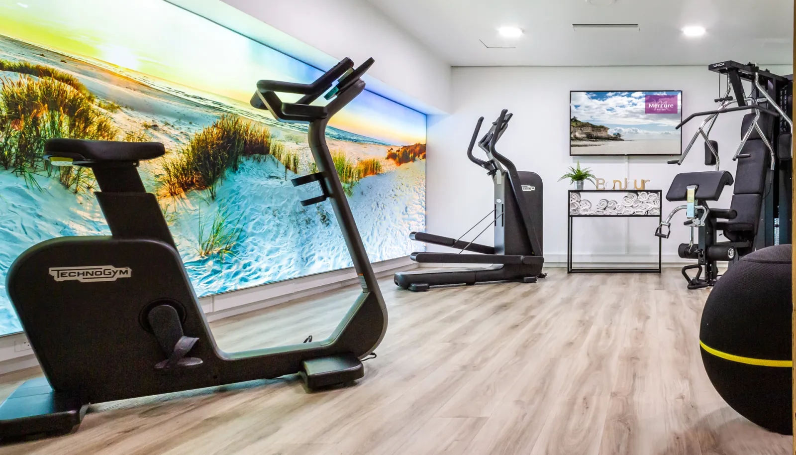 Modern fitness room at Mercure Hotel Bordeaux Centre featuring TechnoGym cardio equipment, large display screen showing a sunny beach scene, and contemporary decor.