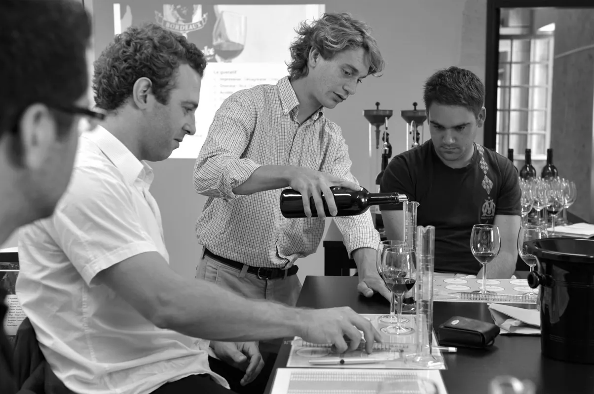 Wine tasting in Bordeaux: a sommelier pours red wine into a glass while three other people watch attentively around a table with glasses and tasting notes.