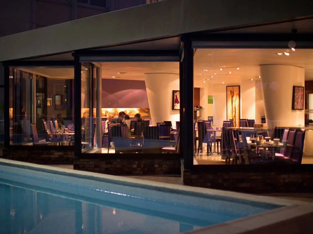 Night view of GourmetBar by Novotel Bordeaux Lac featuring an illuminated glass façade overlooking a swimming pool, with elegantly set dining tables visible inside