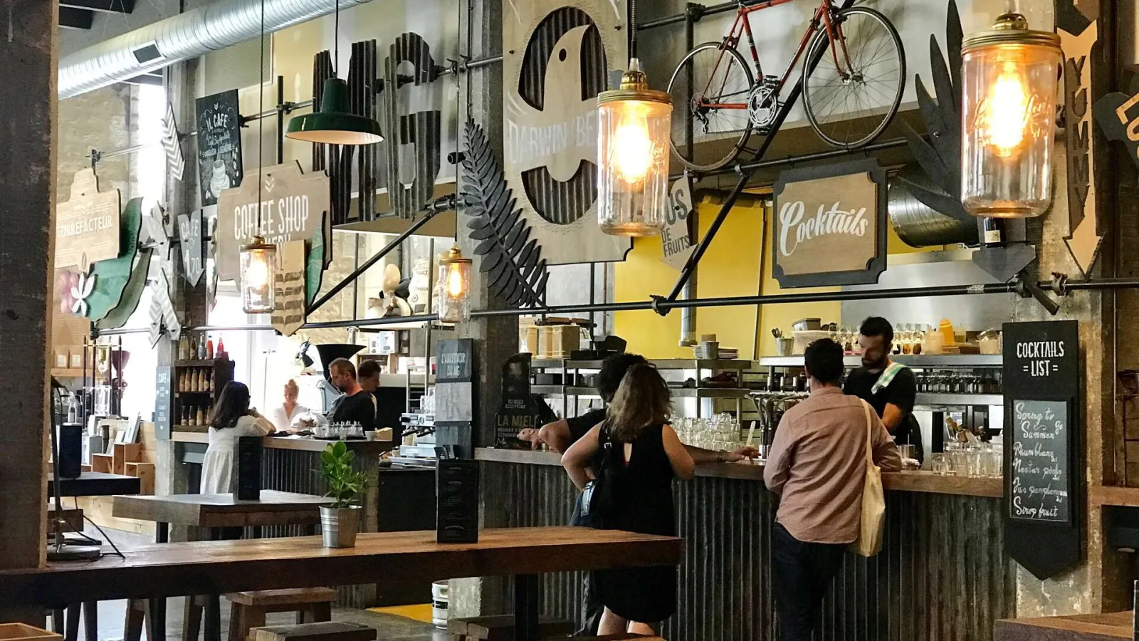 Modern café-bar interior with wooden counter, customers at the bar, industrial décor featuring wall-mounted bicycles, glass pendant lights, and cocktail menu signage in Bordeaux Darwin.
