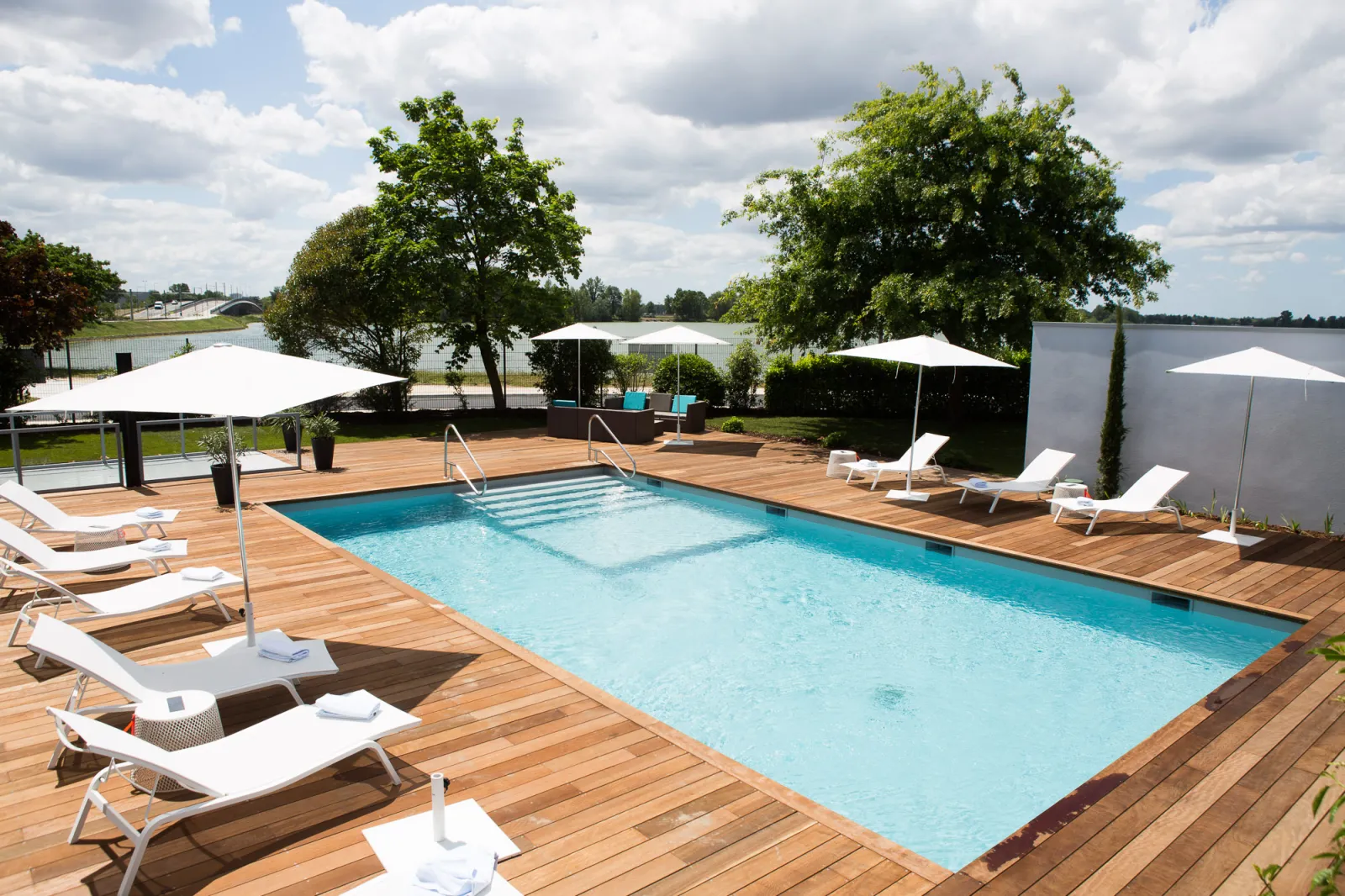 Outdoor swimming pool with turquoise water surrounded by white loungers and umbrellas, wooden deck overlooking Bordeaux Lake