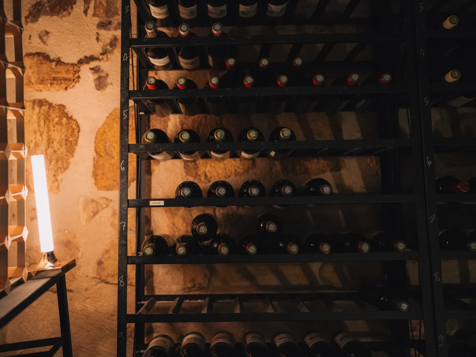 Underground wine cellar with wine bottles stored on black metal shelves, exposed stone walls, and soft lighting.