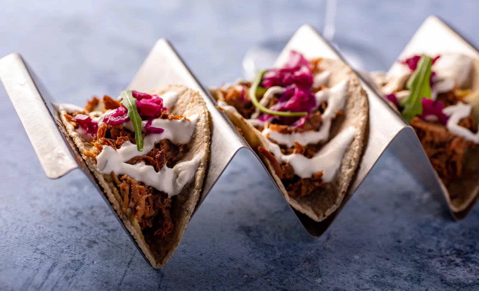Tacos filled with shredded meat, sour cream, red onions and chives, arranged in a row on a gray surface