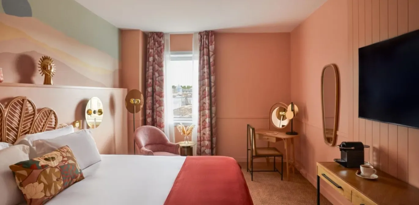 Elegant hotel bedroom with coral walls, white bed with gold palm-shaped headboard, pink armchair, floral curtains, wooden desk and wall-mounted television.