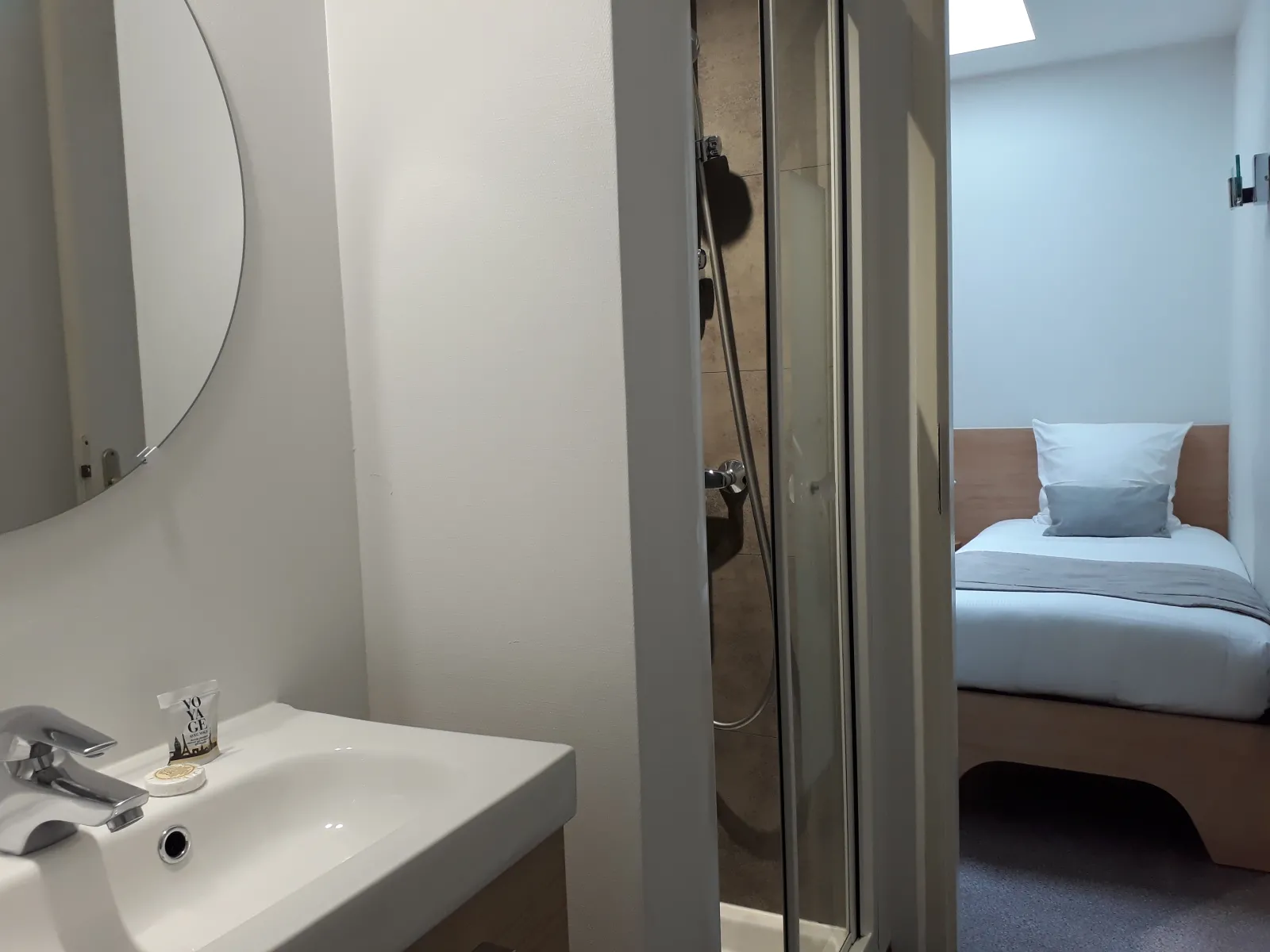 Modern bathroom with white sink and round mirror, opening onto a hotel room with a bed with blue sheets and gray pillows at the Hôtel des Voyageurs in Bordeaux