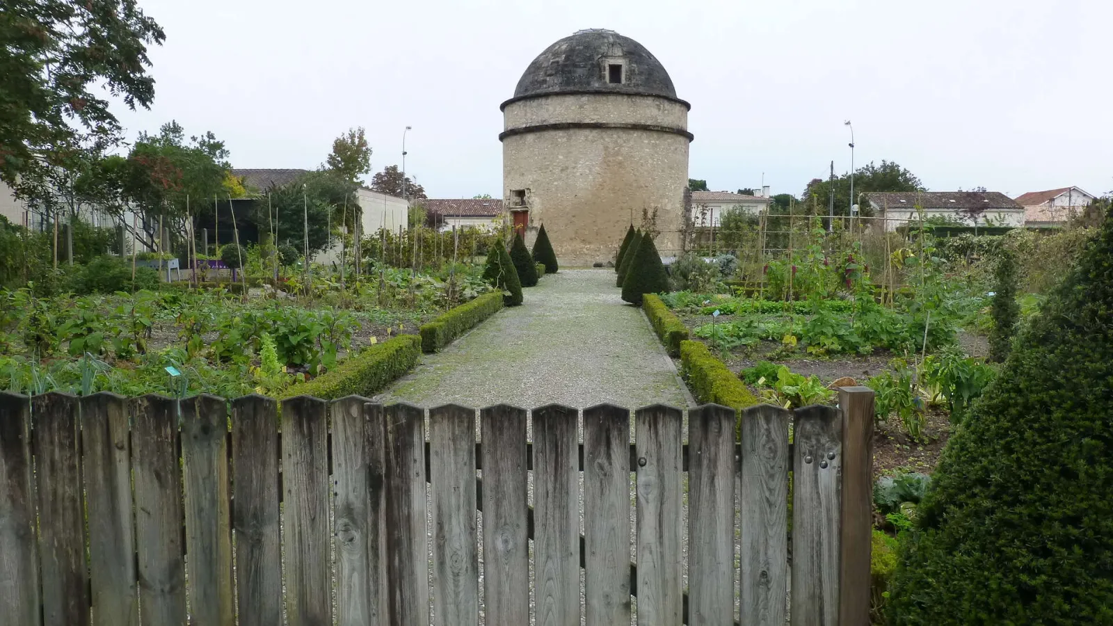 View of the Bordeaux Market Gardening Museum featuring vegetable gardens, a wooden fence in the foreground, and a historic stone building with a dome, surrounded by crops and plants.
