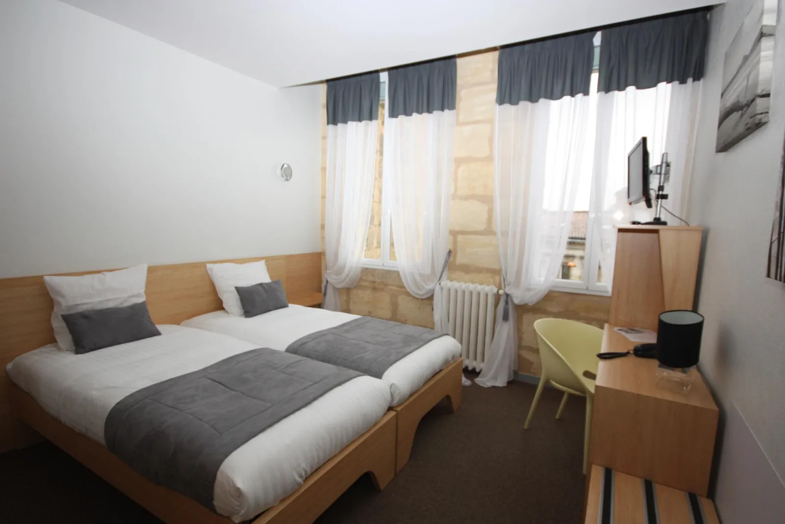 Modern hotel room featuring a light wood double bed with gray and white bedding, wooden desk with yellow chair, windows with gray and white curtains, white radiator, wall-mounted TV, and minimalist décor.