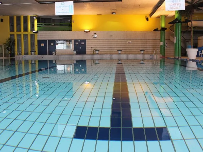 Empty Olympic swimming pool at Pessac Aquatic Stadium in Bordeaux, featuring light blue tiled basin with black and white lane lines, yellow and green walls, diving equipment, and spectator areas.