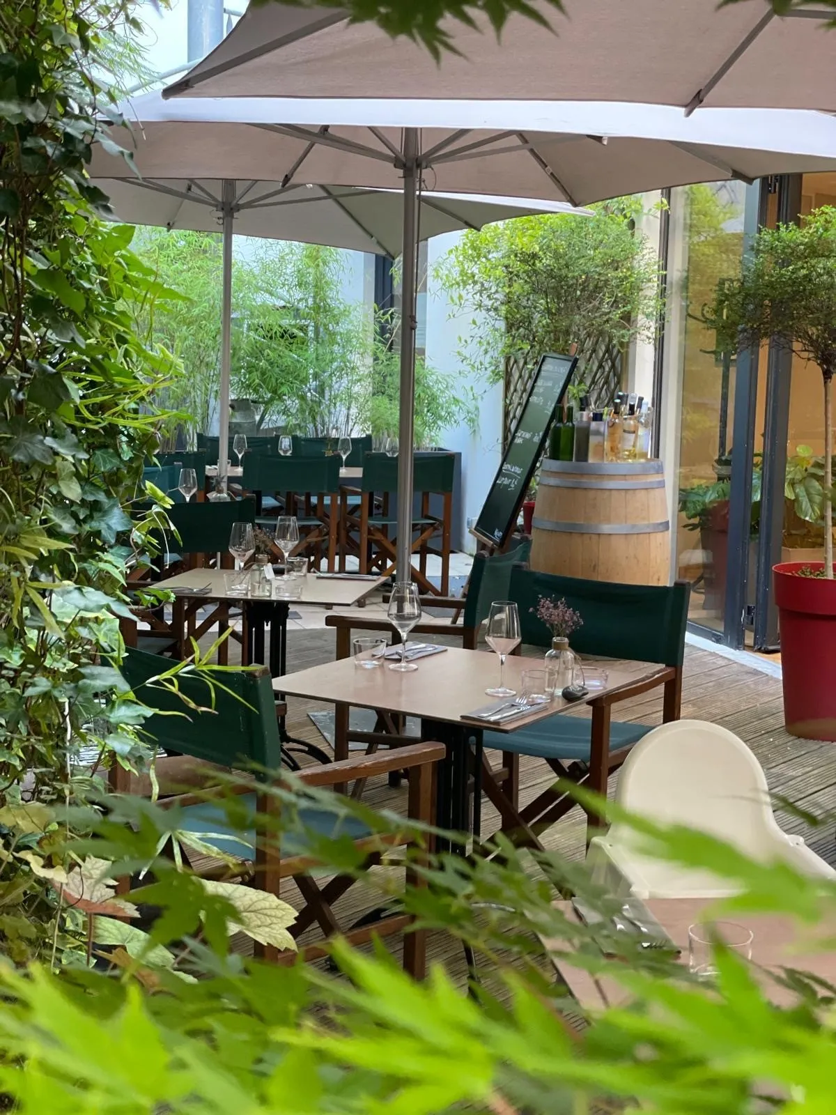 Shaded outdoor dining terrace at Les Tables Vatel restaurant in Bordeaux featuring wooden tables, beige umbrellas, lush greenery, and a wine barrel in the background