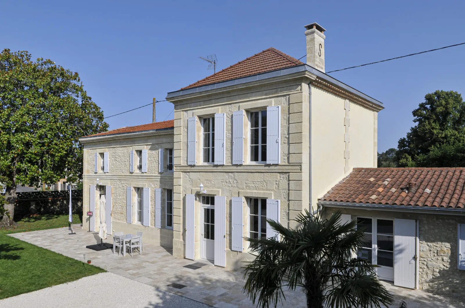 Façade of Château d'Osmond in Bordeaux, a white stone building with blue shutters, red tile roof and a square tower, surrounded by trees and greenery.
