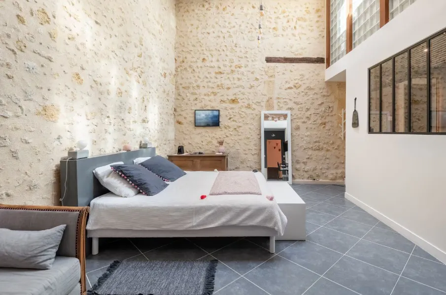 Spacious bedroom with exposed stone walls, modern gray bed with white linens, wooden furniture, gray tiled flooring, and mezzanine with windows at Gatto Verde in Bordeaux