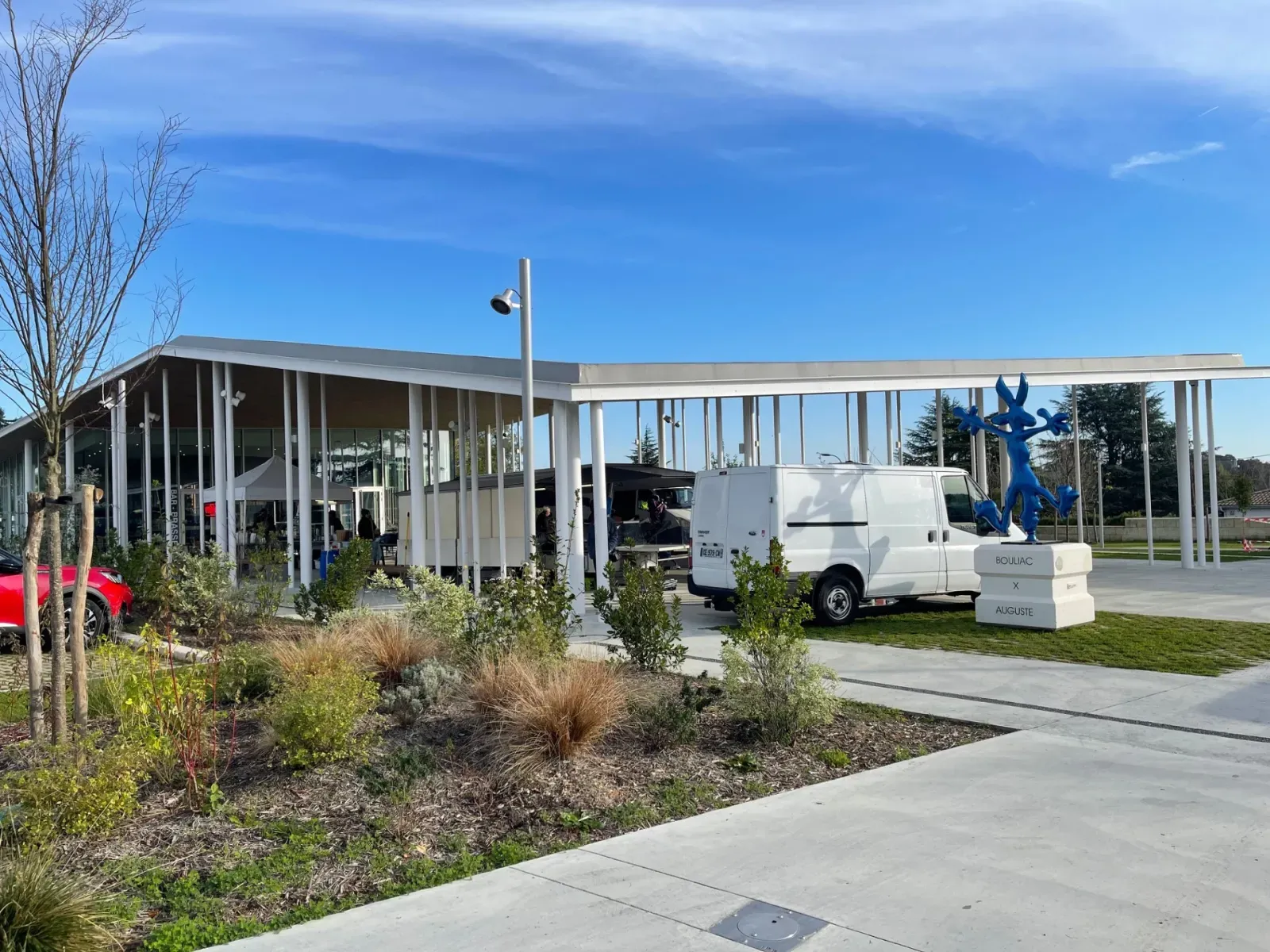 Vettiner Hall at Bouliac Market in Bordeaux, modern building with white columns, glazed canopy, blue statues and parked white van under clear sky