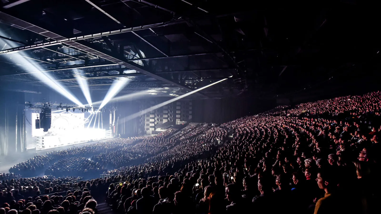 Panoramic view of the Arkea Arena concert hall in Floirac, Bordeaux, showing a large crowd of spectators facing an illuminated stage with blue spotlight beams.