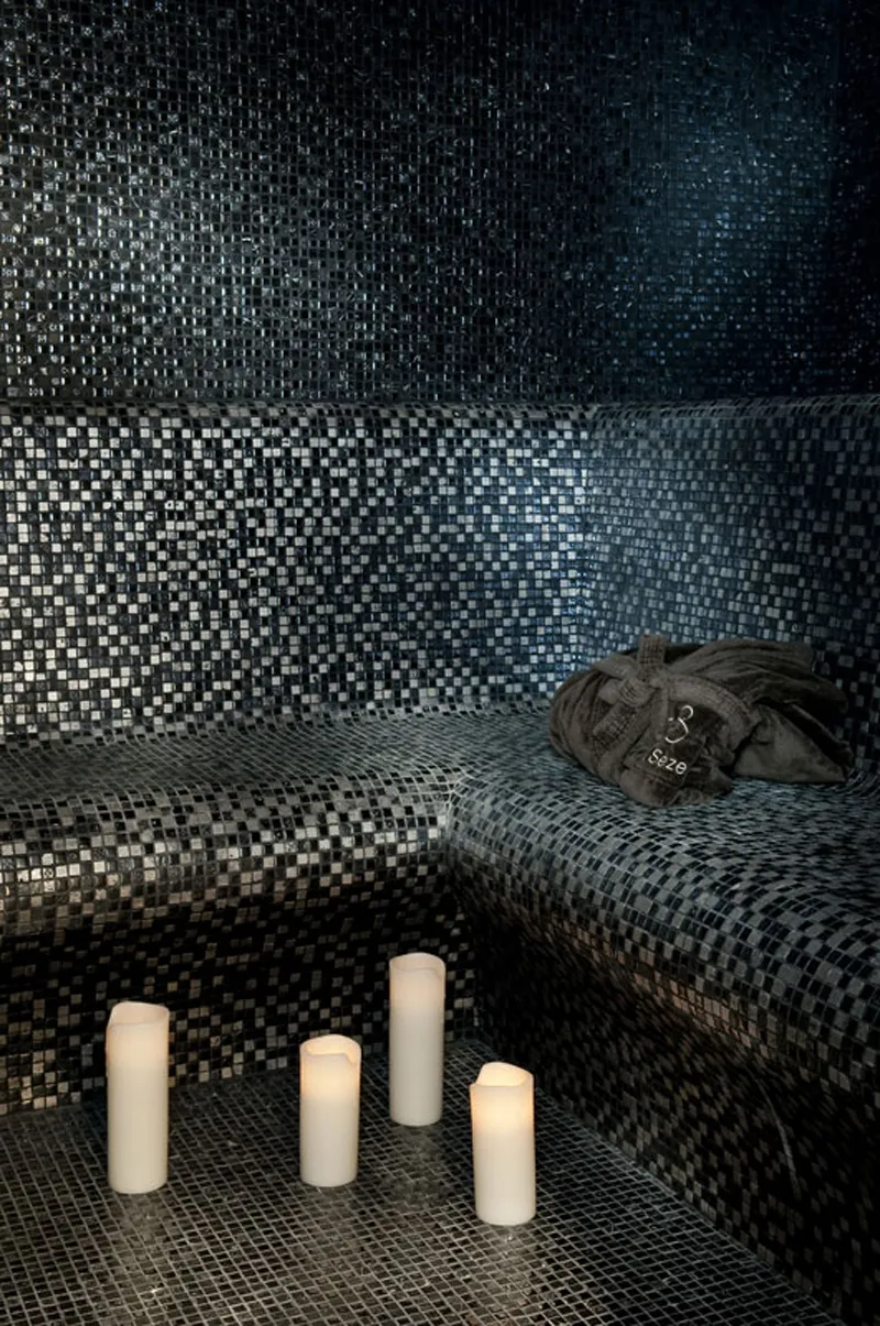 Spa space with gray and white mosaic tiles, four lit candles on the floor, and a black towel on a stone bench