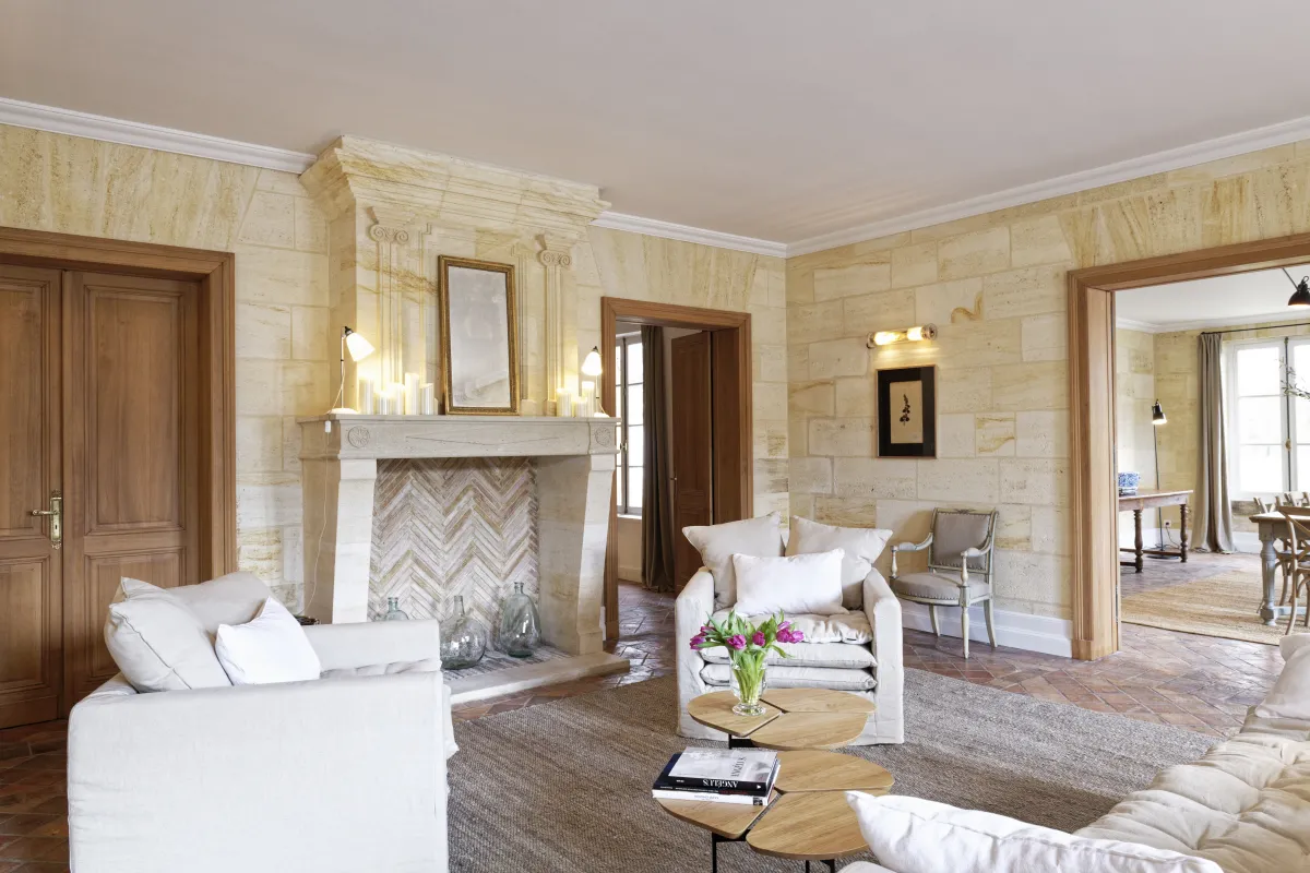 Elegant living room at Maison La Fleur de Boüard in Bordeaux featuring a stone fireplace with herringbone pattern, white upholstered sofas, light stone walls, wooden doors, and multiple room connections