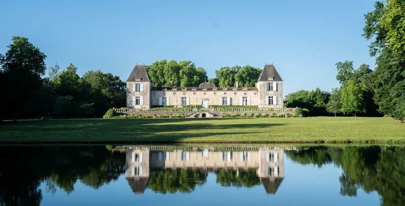 Château de Sales near Bordeaux, a classical stone mansion with two conical turrets, perfectly reflected in a calm pond, surrounded by lush greenery.