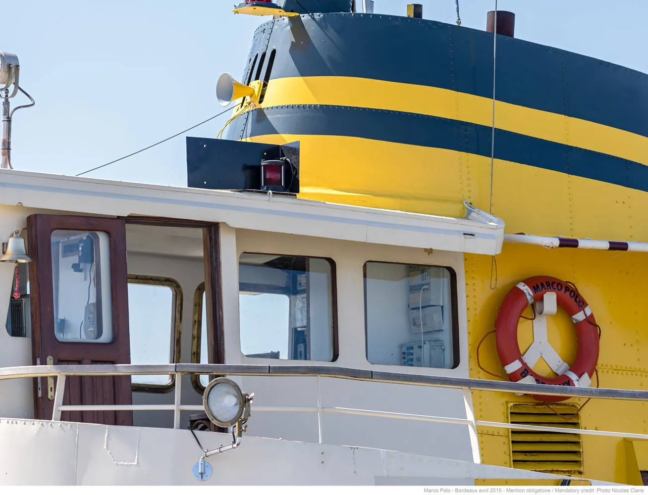 Close-up of a yellow and blue river cruise ship docked in Bordeaux, featuring the wheelhouse, portholes, a red and white life ring, and navigation equipment.