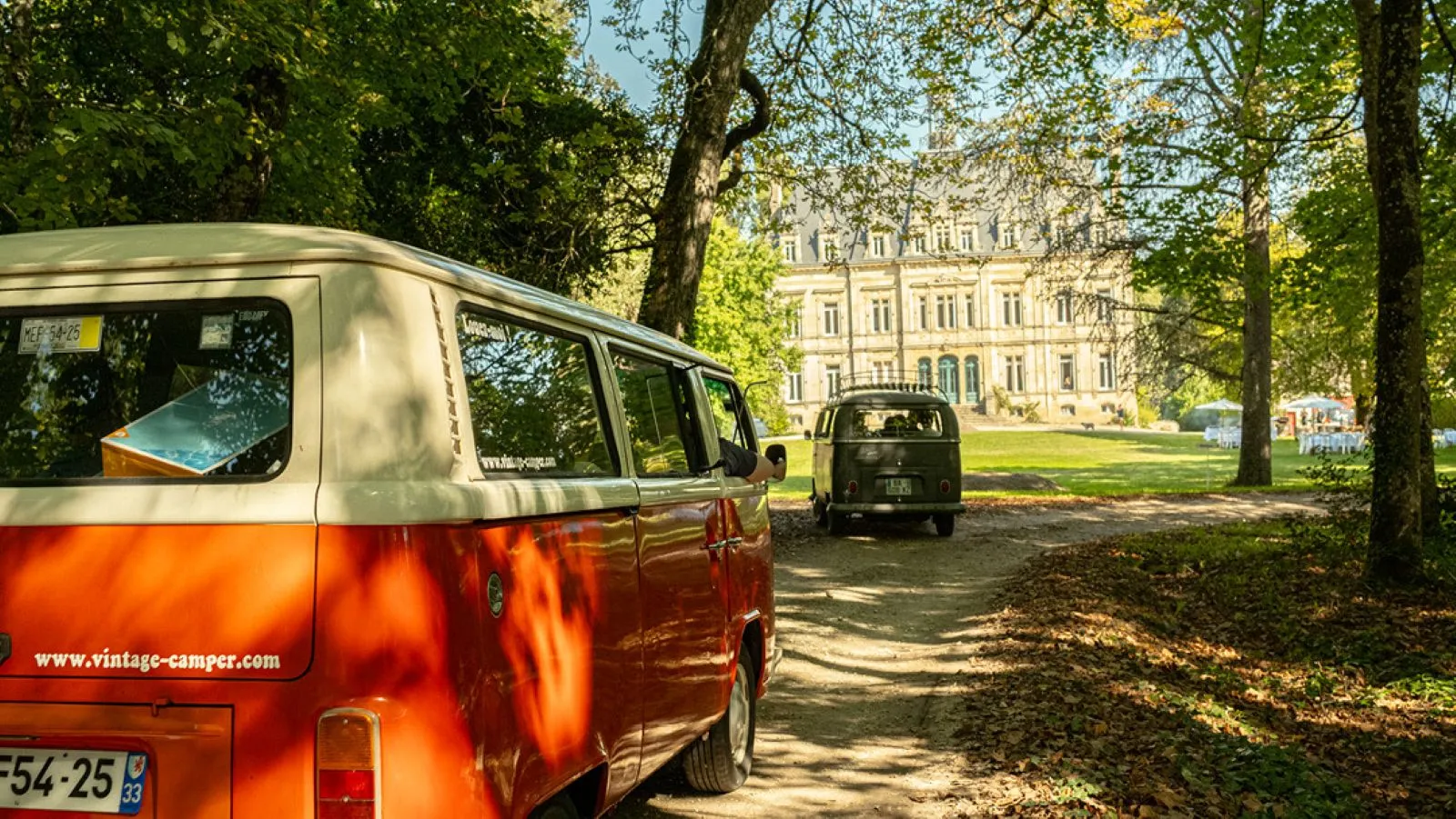 Two vintage camper vans parked under trees in a park in Bordeaux, with a classical building in the background