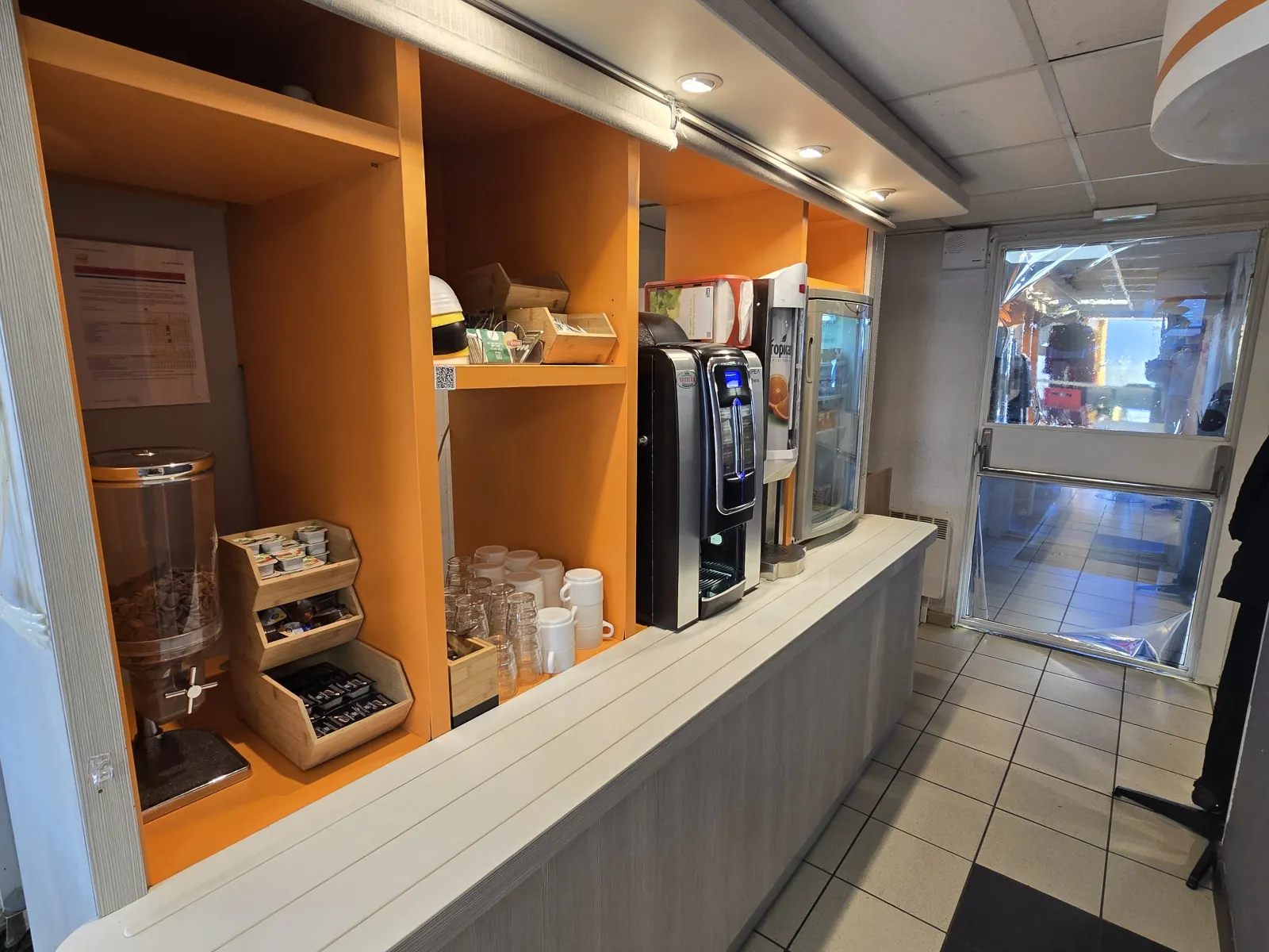 Coffee and breakfast area of Hôtel Première Classe Bordeaux Ouest Eysines featuring beverage dispensers, orange shelving units, service equipment and access to the terrace.