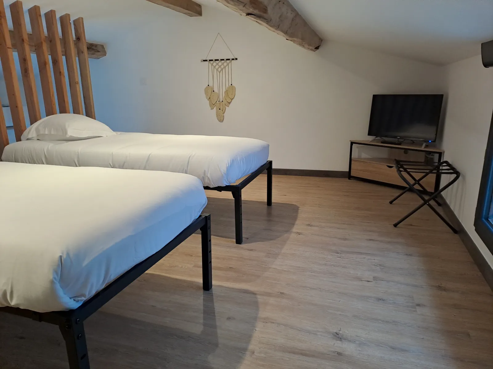 Bedroom at Anthocyane guesthouse in Château Pierre de Montignac near Bordeaux, featuring two single beds with white linens, wooden headboard, macramé wall decoration, TV, and exposed ceiling beams.