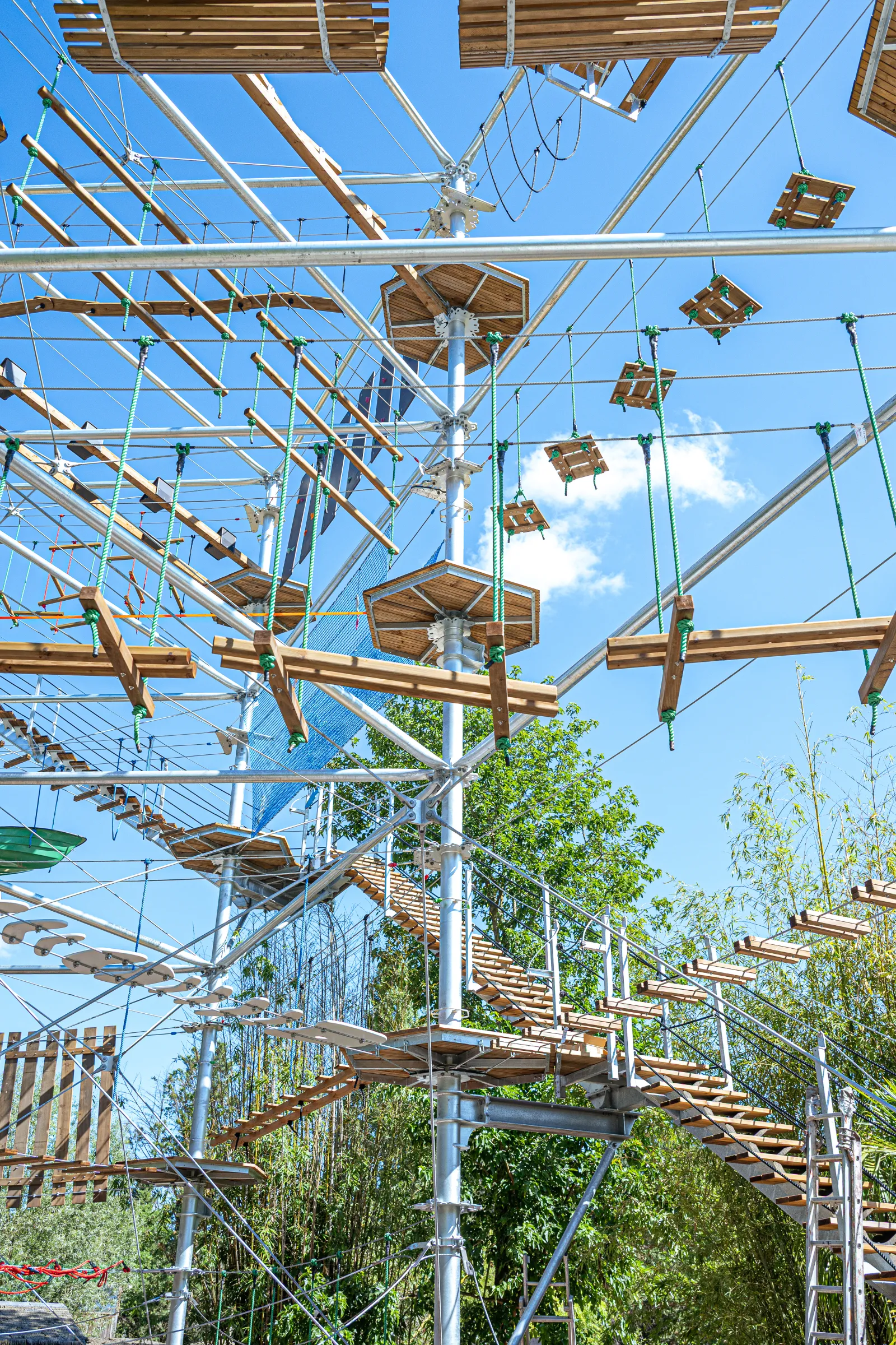 Adventure park The Cimes of Bordeaux in Pessac featuring suspended aerial course, rope bridges, wooden beams and safety nets among the trees under blue sky.
