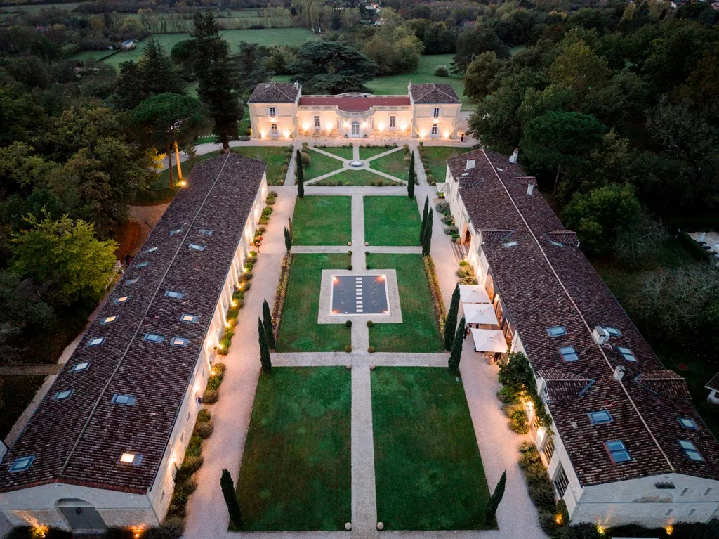Aerial view of a Bordeaux château with symmetrical French formal gardens, flanked by two lateral wings, surrounded by forests and green meadows.