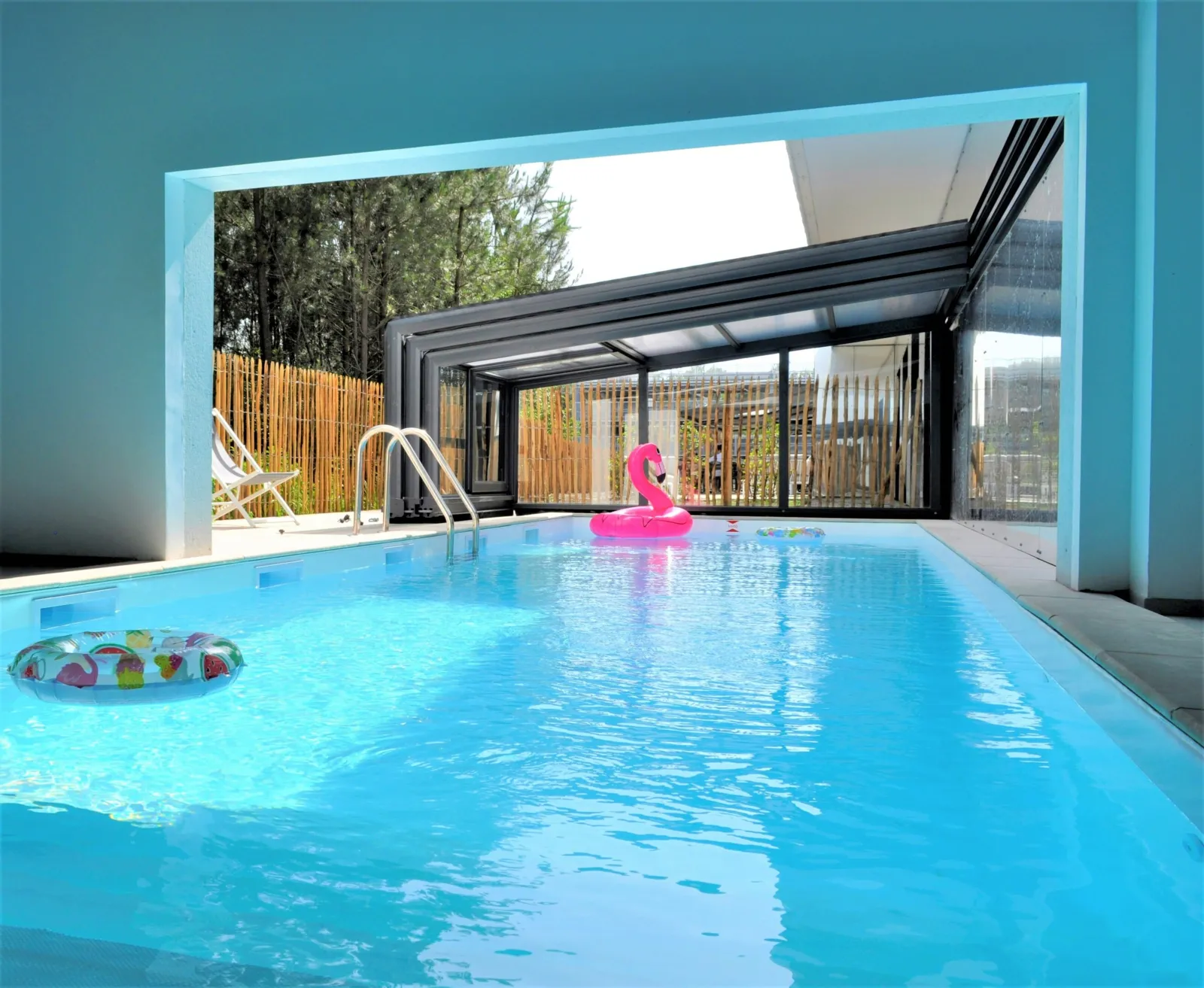 Indoor heated swimming pool at Holiday Inn Bordeaux Mérignac featuring crystal-clear blue water, white entry stairs, pink flamingo float, and large windows overlooking an outdoor terrace with trees.