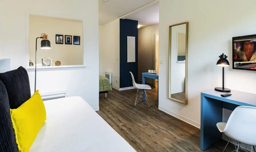 Modern studio apartment at Ténéo Aparthotel in Bordeaux Saint-Jean featuring a white bed with yellow pillow, blue desk, workspace chair, black partition wall, and wooden flooring.