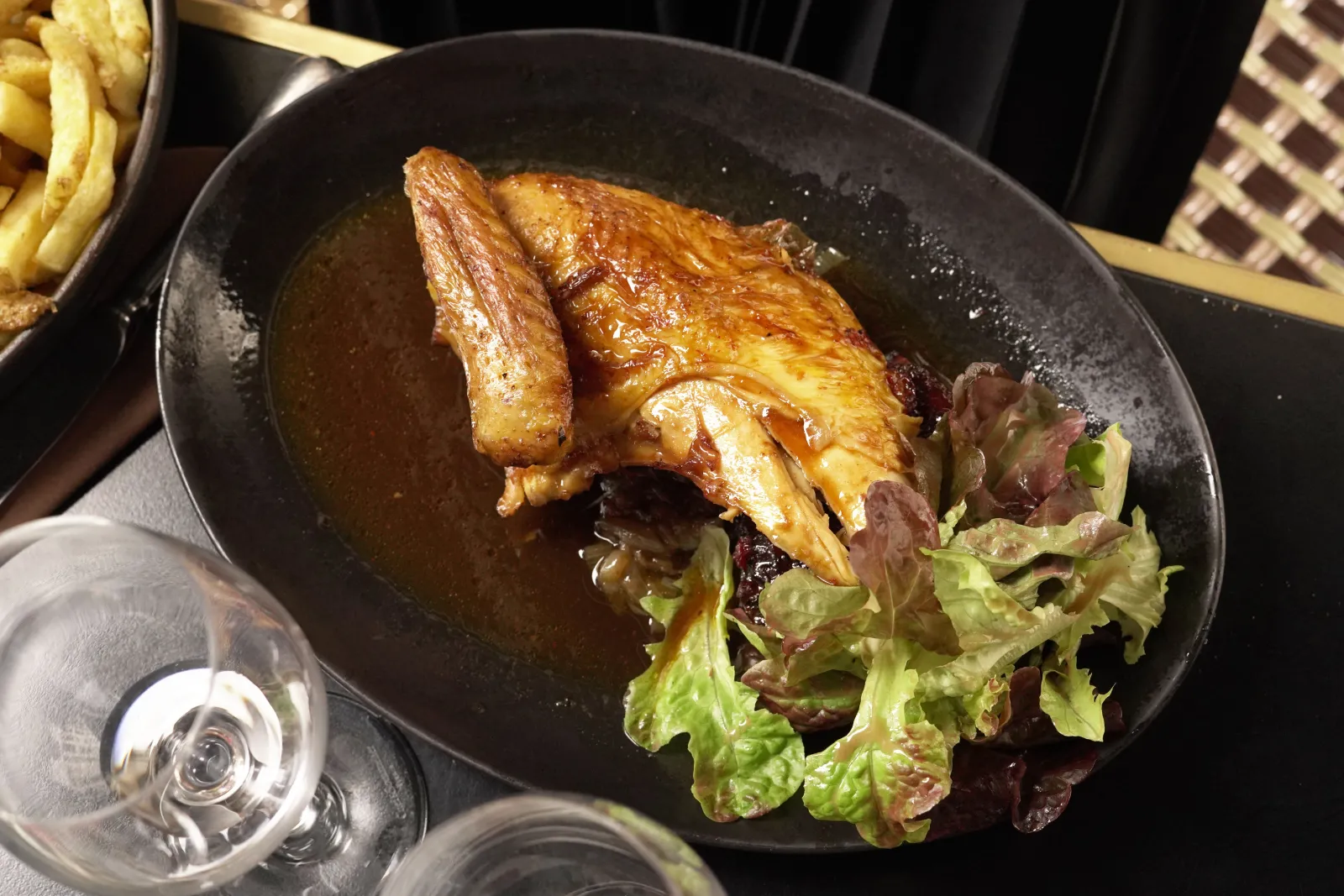 Golden roasted duck served in a black plate with green salad and sauce, accompanied by fries and a glass of white wine