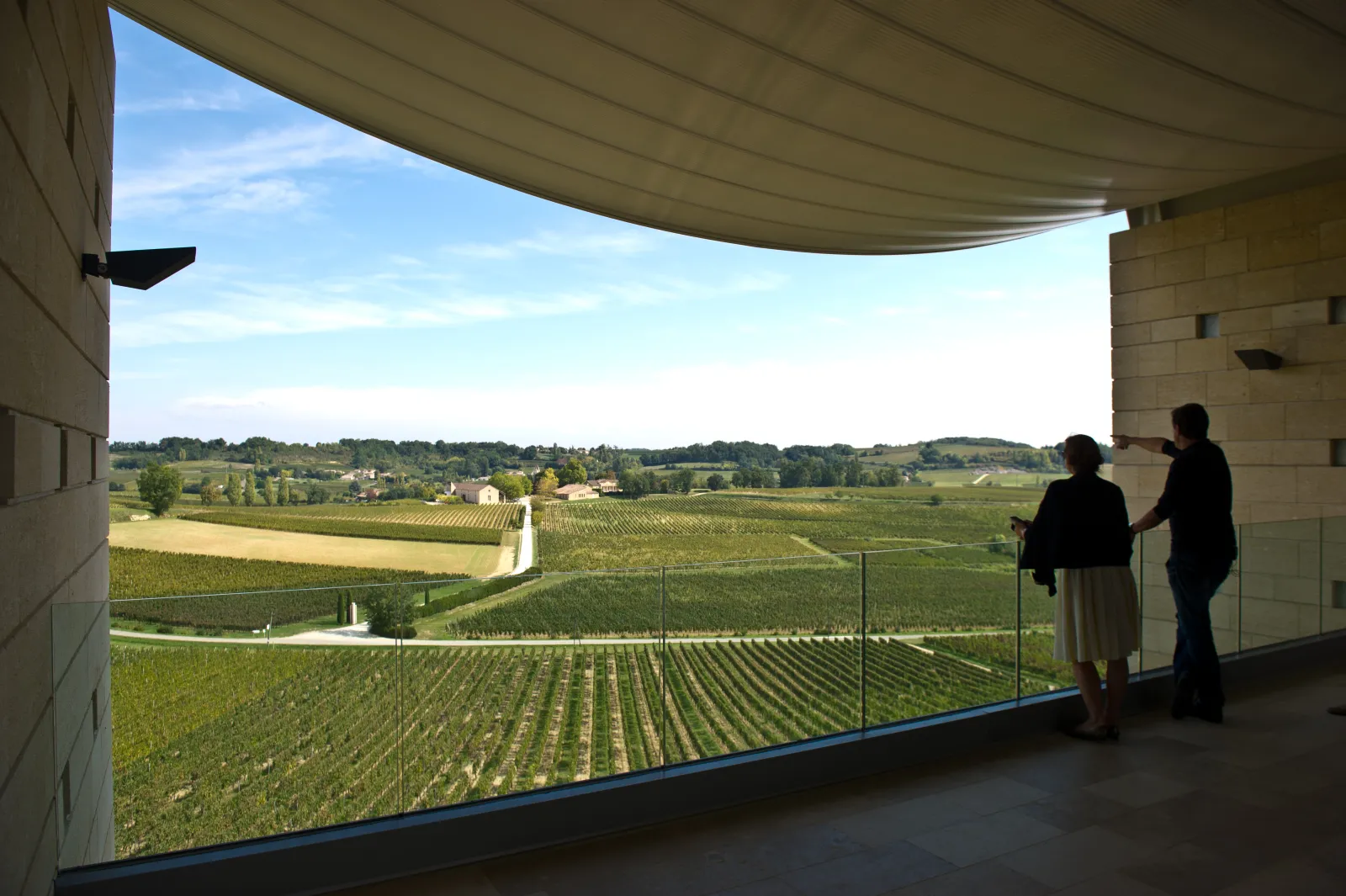 Panoramic view from a covered terrace at Château Faugères overlooking Bordeaux vineyards, with two visitors admiring the vine rows and rolling landscape