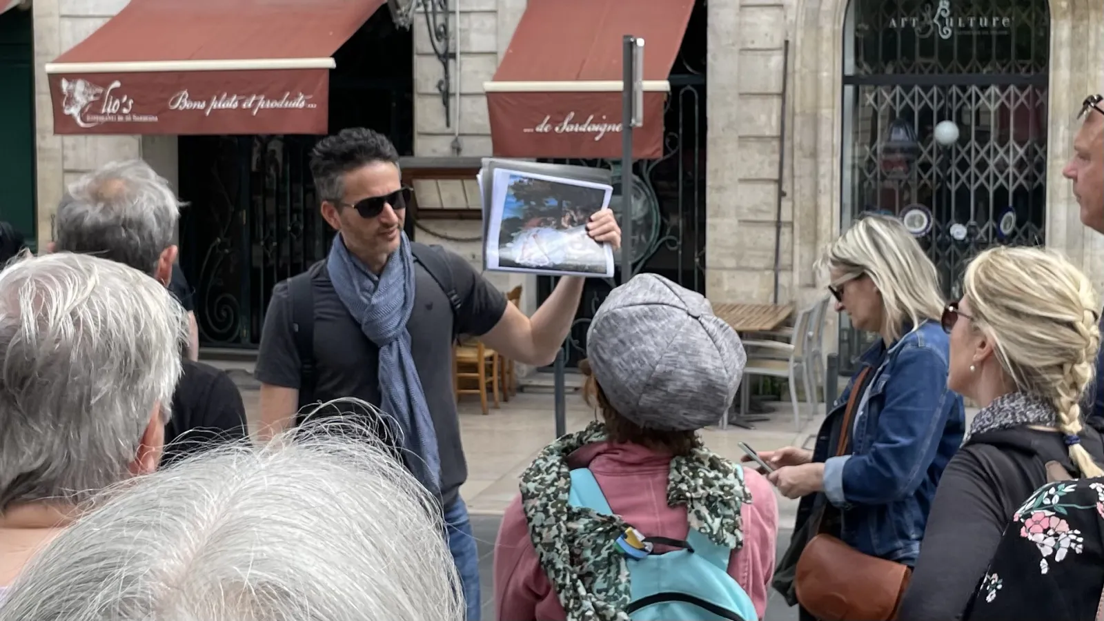 A tour guide shows a tablet to a group of visitors on a Bordeaux street, in front of historic buildings and burgundy awnings.