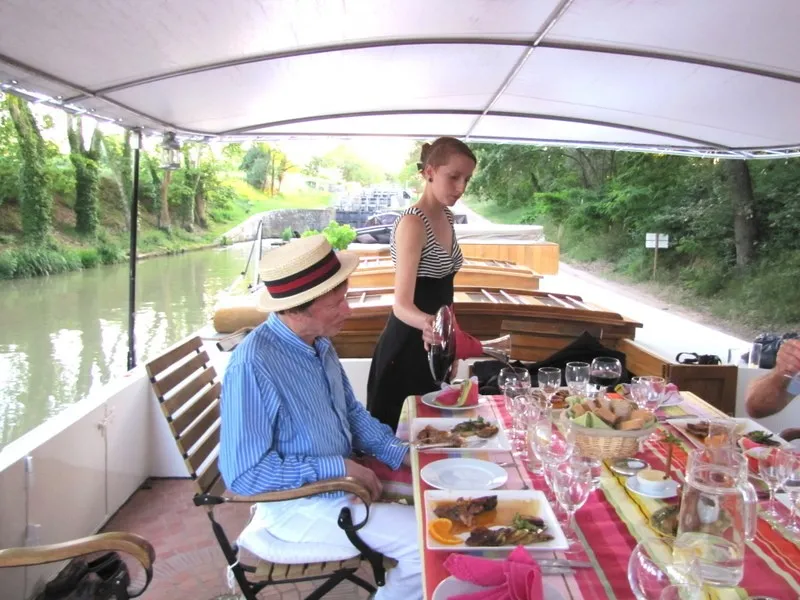 Gourmet meal on a barge along the Garonne River in Bordeaux, featuring a man in a blue shirt and straw hat seated at a decorated table, a standing woman in the background, under a white canopy, with green trees and buildings visible.