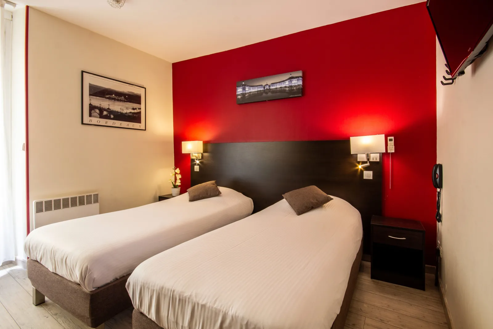 Hotel room with two single beds with white linens, black headboard, red and beige walls, gold bedside lamps, and framed photographs at Hotel Notre Dame in Bordeaux.