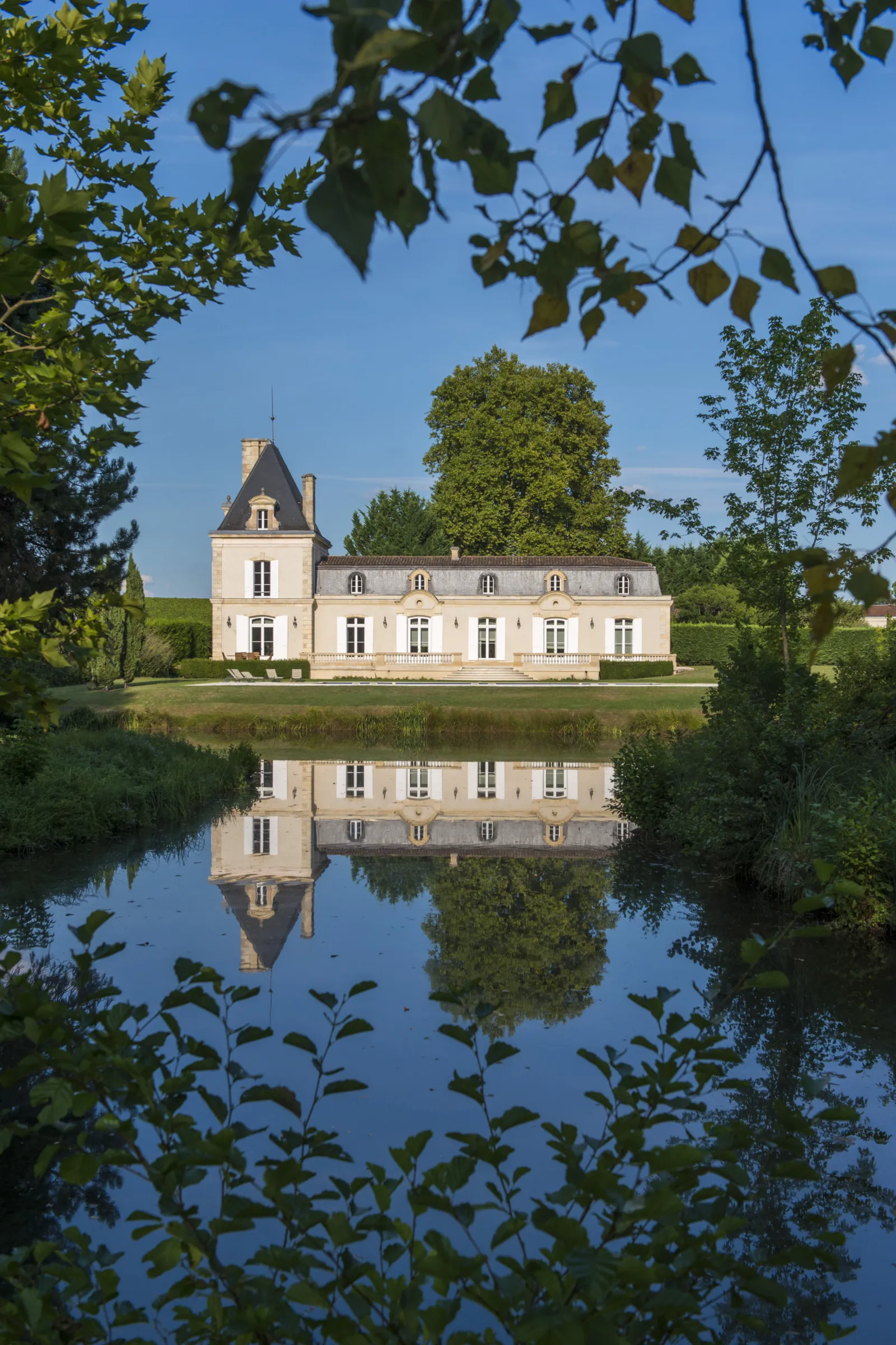 Château Larrivet Haut-Brion in Bordeaux, an elegant 19th-century white mansion with mansard roof, perfectly reflected in the surrounding water basin, surrounded by lush greenery.