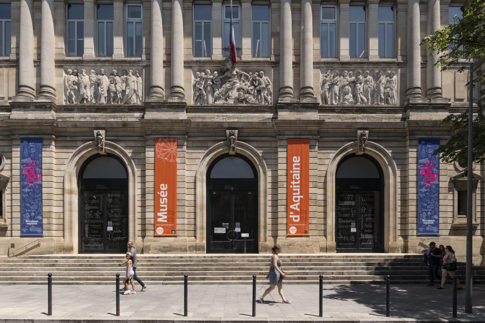 Facade of the Museum of Aquitaine in Bordeaux featuring tall columns, sculptural friezes, orange and blue banners, three arched doors, and visitors in the square.
